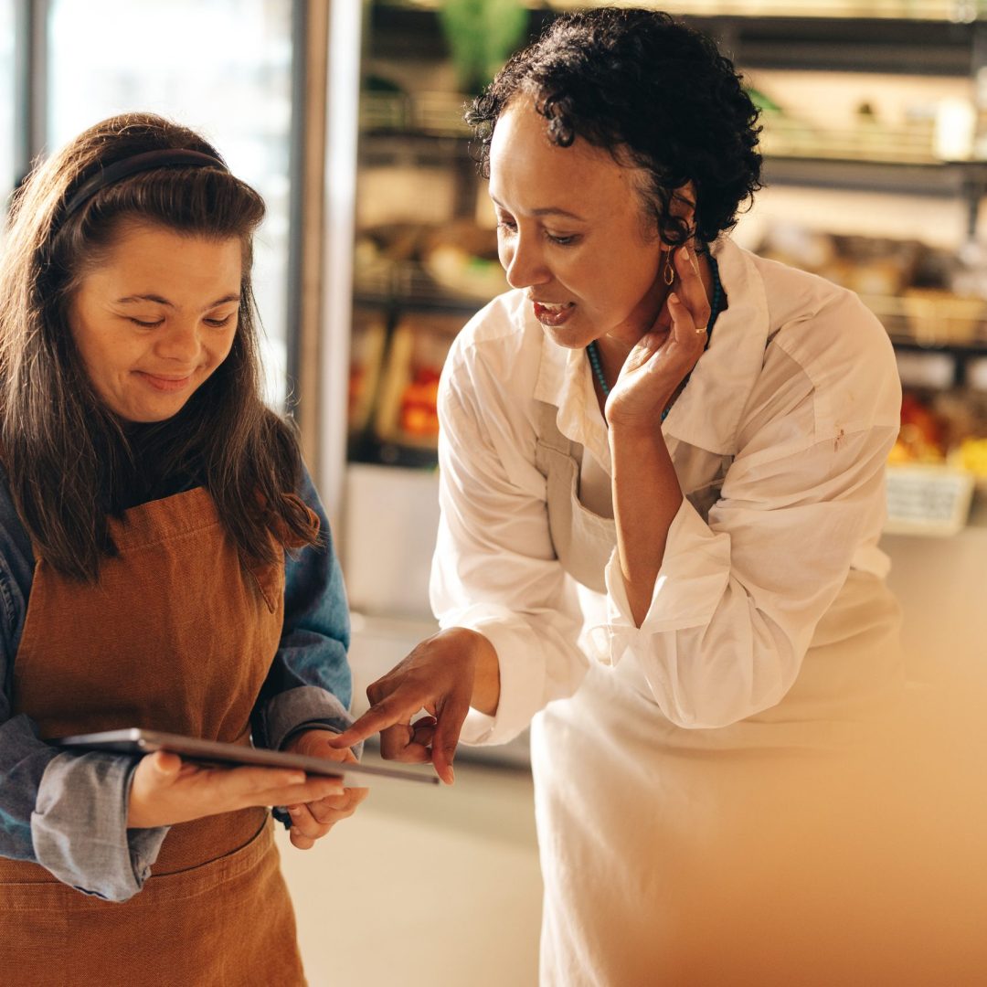 Two supermarket workers managing online grocery orders on a digital tablet. Empowered woman with Down syndrome running a successful small business with her female colleague.